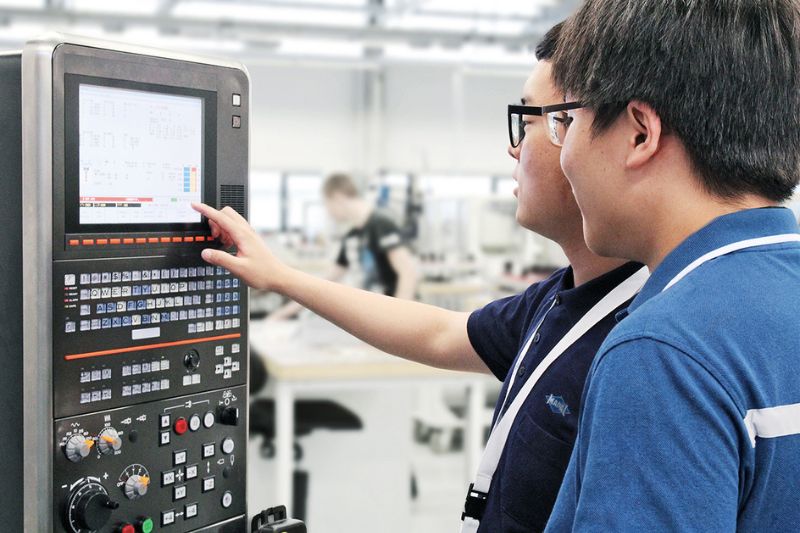 The image shows two MAPAL employees in Korea programming a machine.